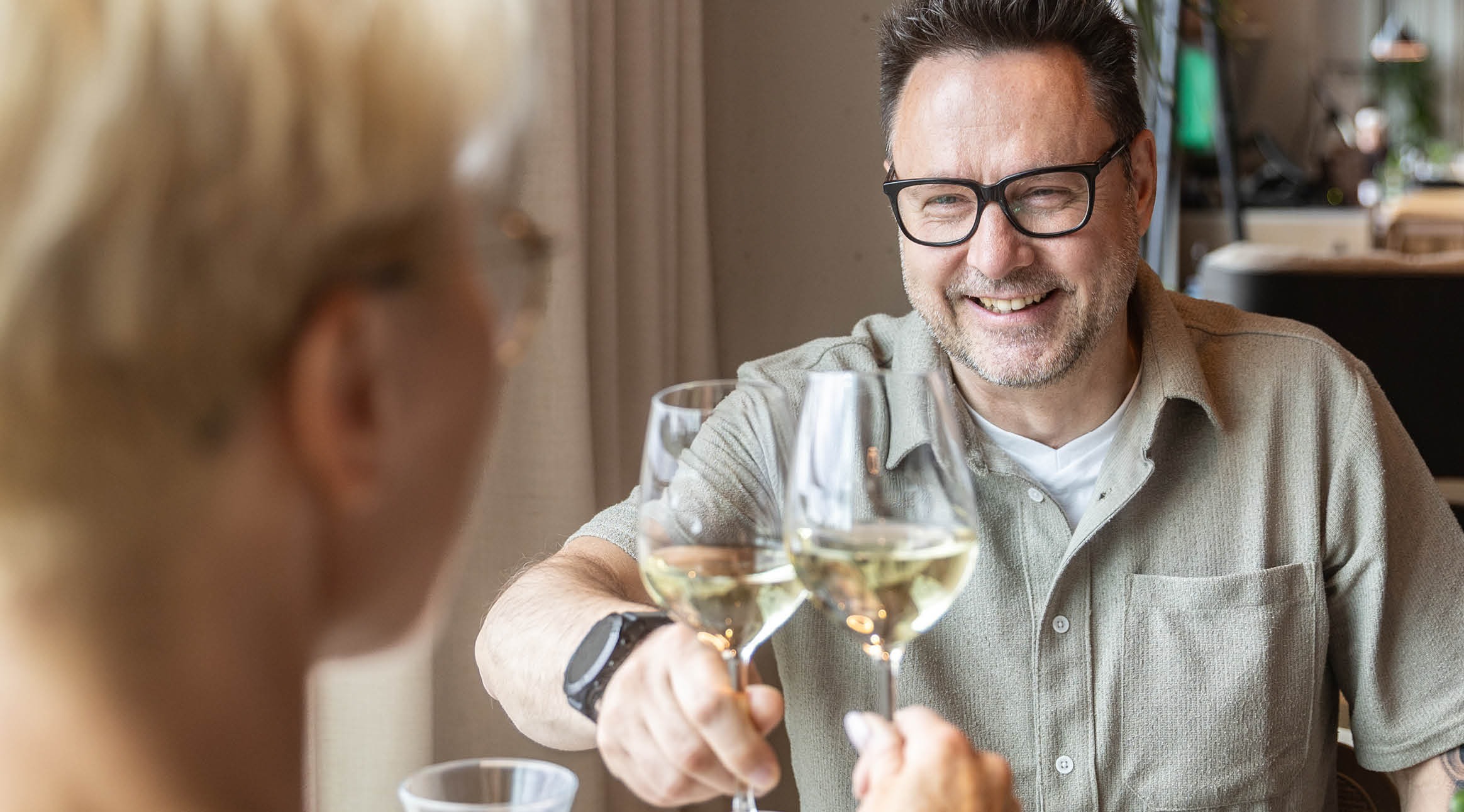 Two people toasting with wine glasses in a restaurant. Close-up of a smiling person wearing glasses and a light-colored shirt.
