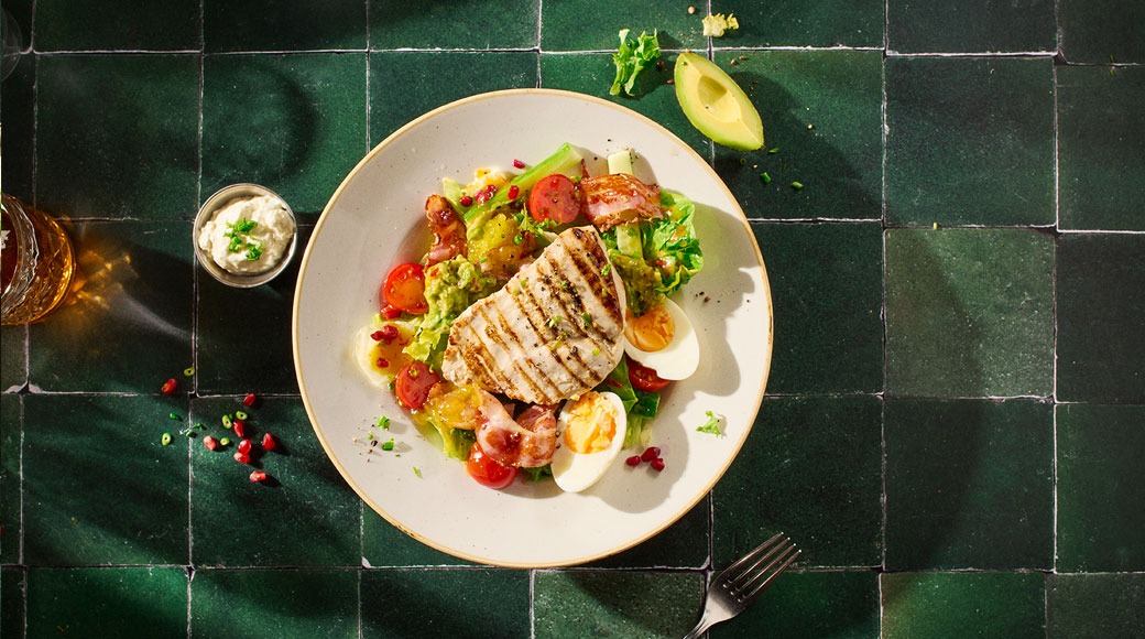 Kulhollinen salaattia, jonka päällä grillattu kanafilee, kananmunaa, pekonia ja kirsikkatomaatteja. Vieressä dippikuppi, avokado ja haarukka vihreällä laattapöydällä.