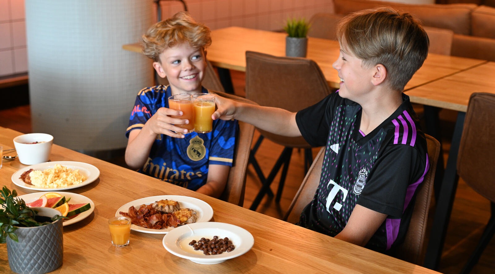 Two children toasting with orange juice at a restaurant table. Various breakfast foods are served on the table.