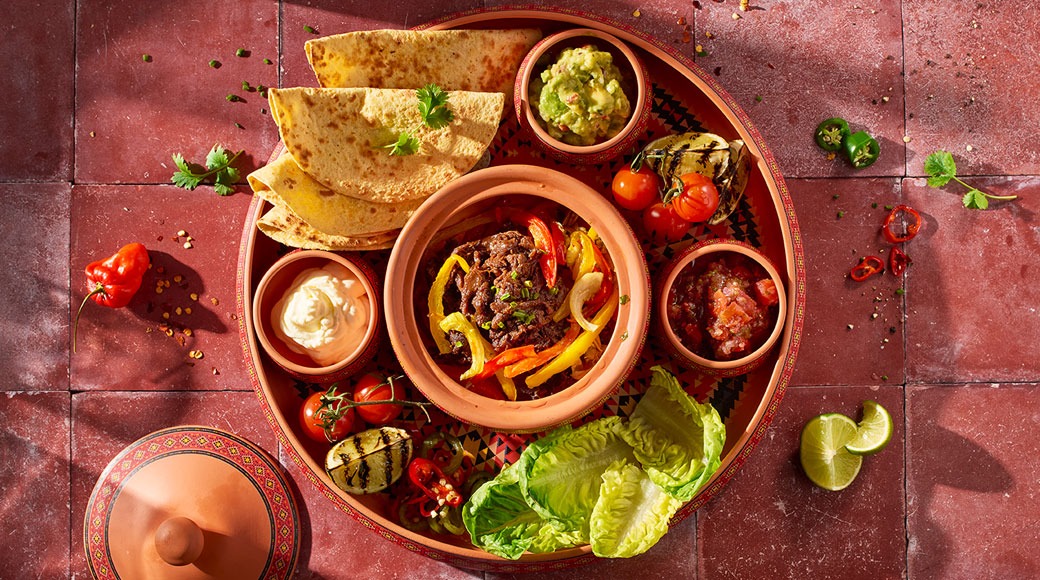 Pyöreä tarjotin, jolla on fajita-lihaa ja paprikaa, tortillat, guacamole, smetana, salsa, salaatinlehtiä ja grillattua kasvista punaisella laatalla.