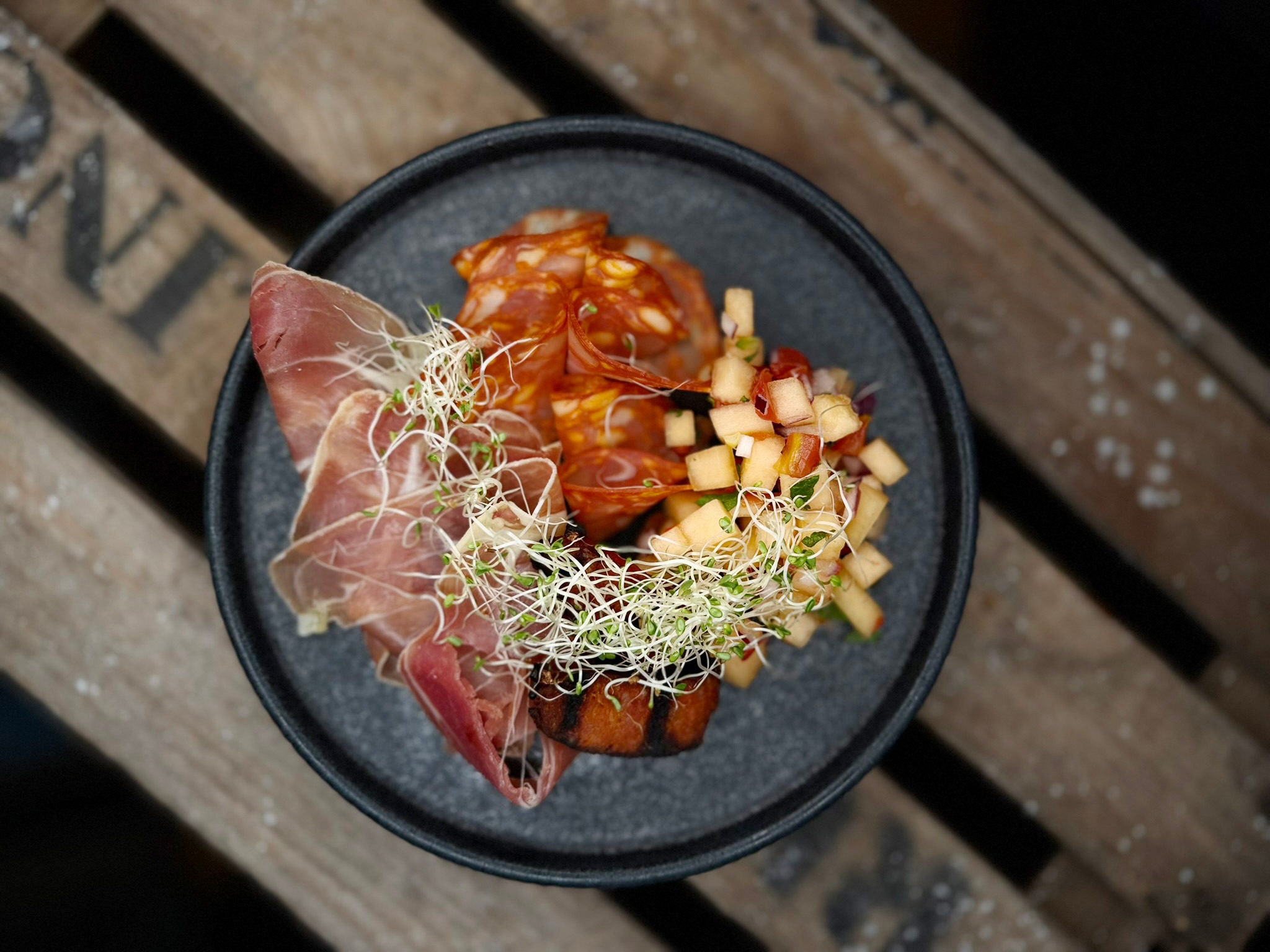 Dark plate featuring a charcuterie dish with prosciutto, salami, diced fruit and microgreens, placed on a wooden table.