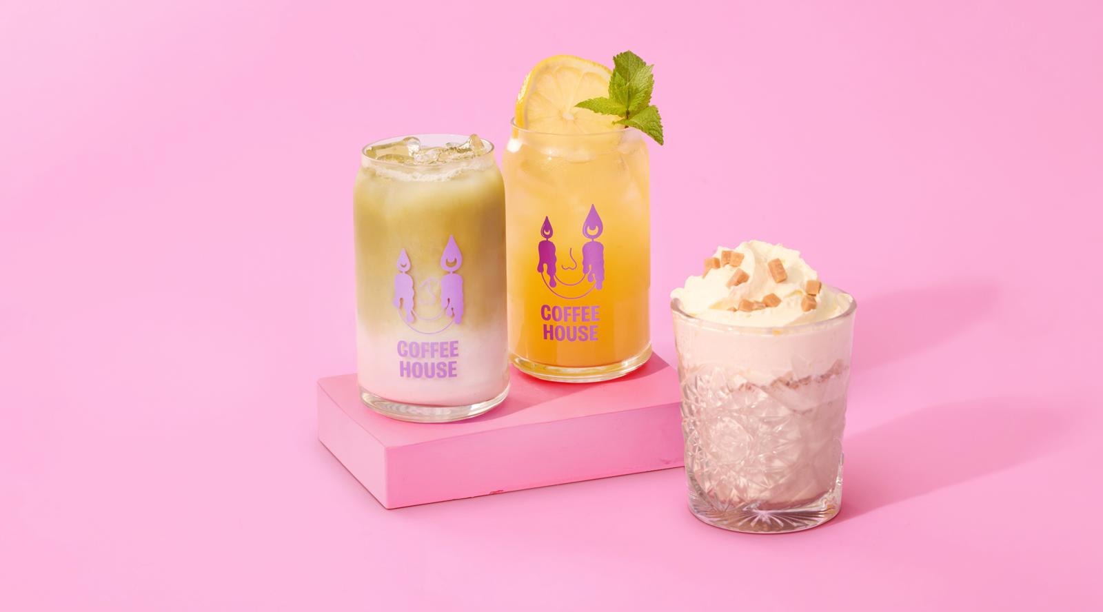 Three different drinks on a pink background: a milky coffee, an iced drink garnished with lemon and mint, and a frosty beverage topped with whipped cream.