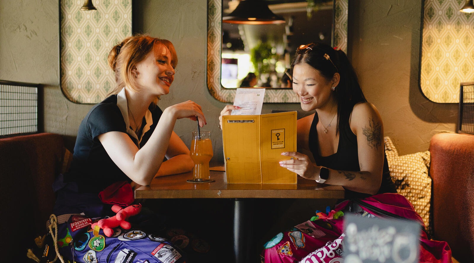 Two smiling women sit at a table on upholstered bench seats, looking at a menu. A drink is on the table. The space has warm lighting.