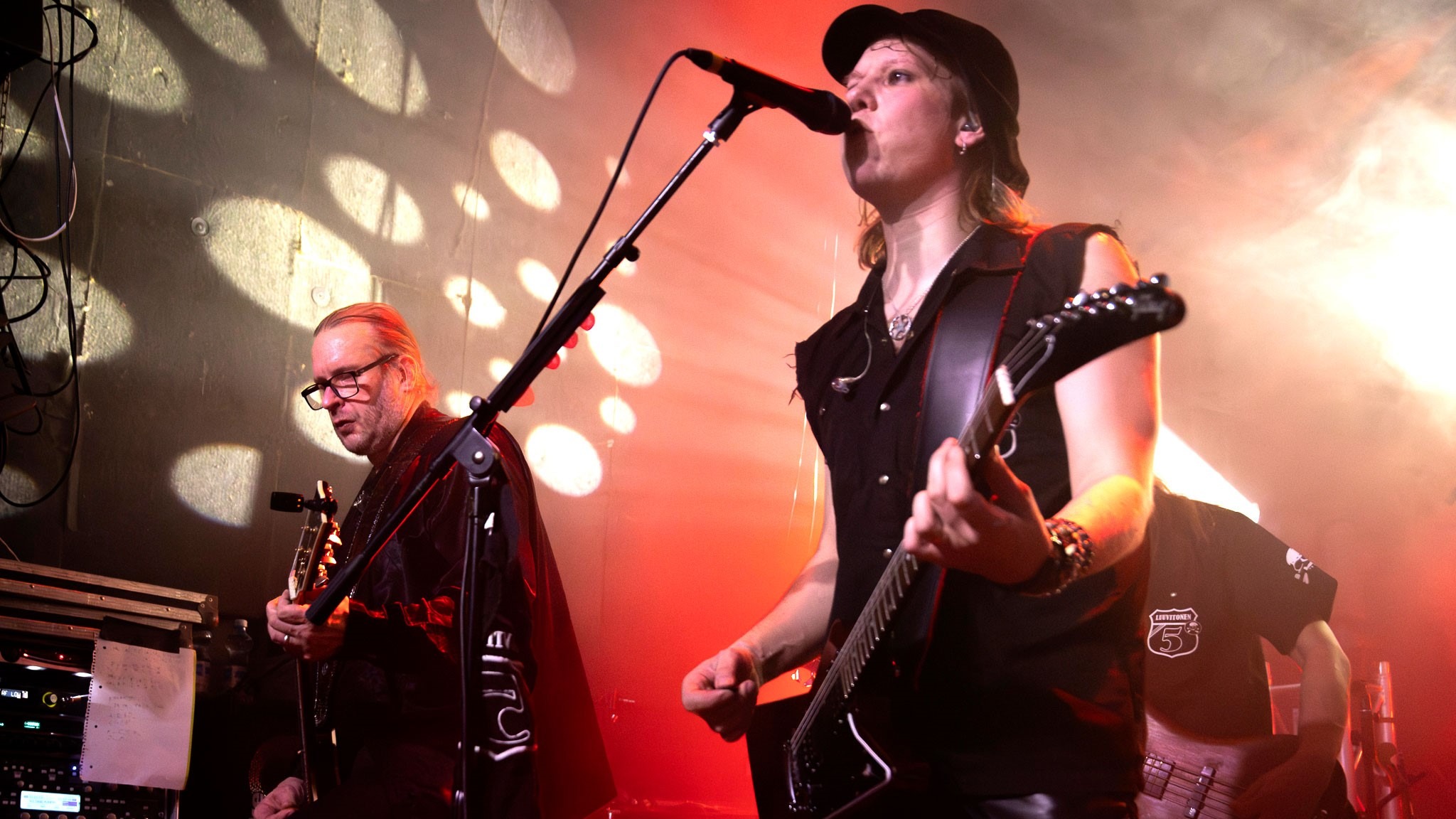 Band uppträder på scen, i förgrunden sångare med gitarr vid mikrofonen och en annan musiker som spelar bas. Rödaktig belysning skapar stämning.