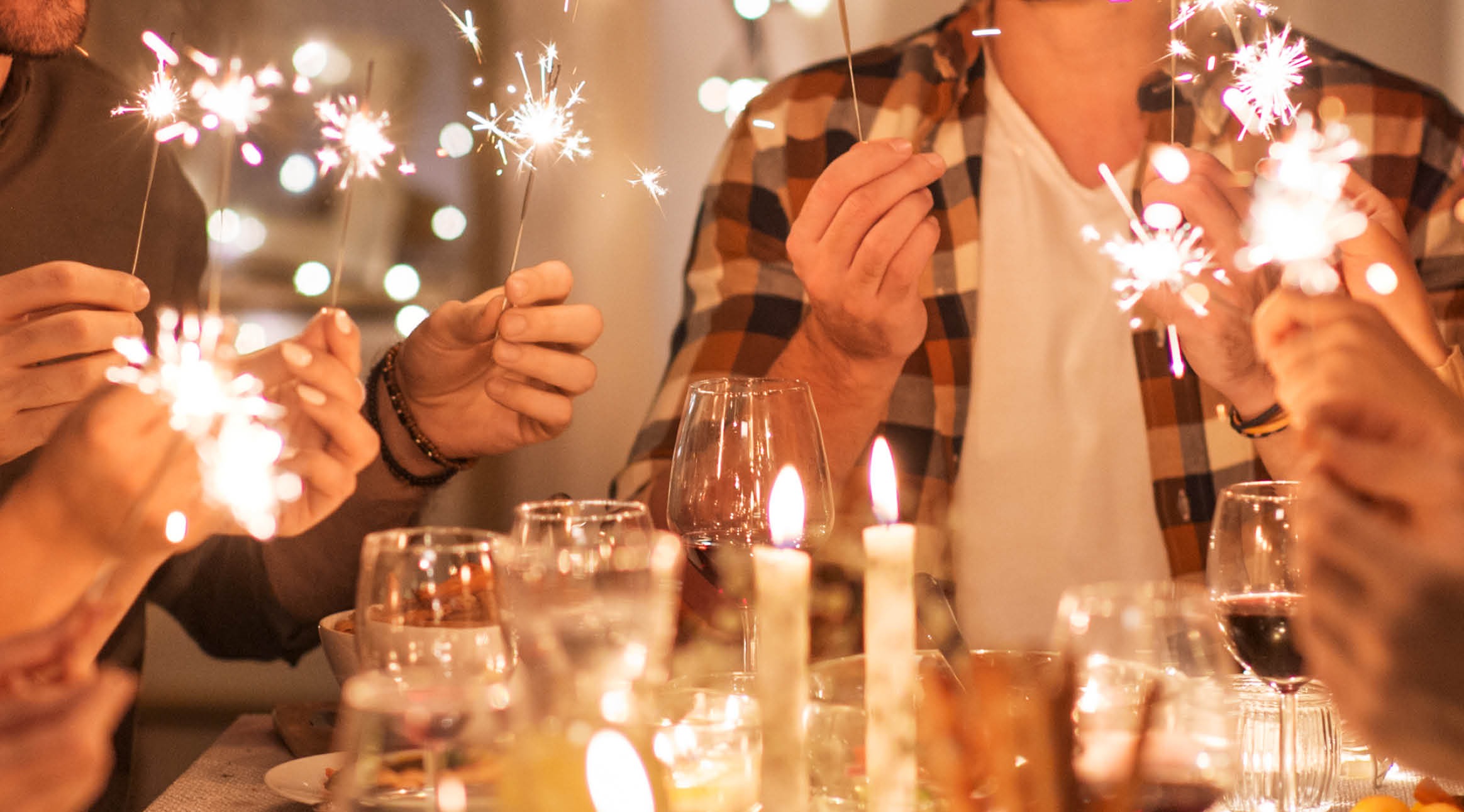 Ihmiset pitävät palavia tähtisädetikkuja juhlapöydän ääressä. Pöydällä on viinilaseja ja palavia kynttilöitä.