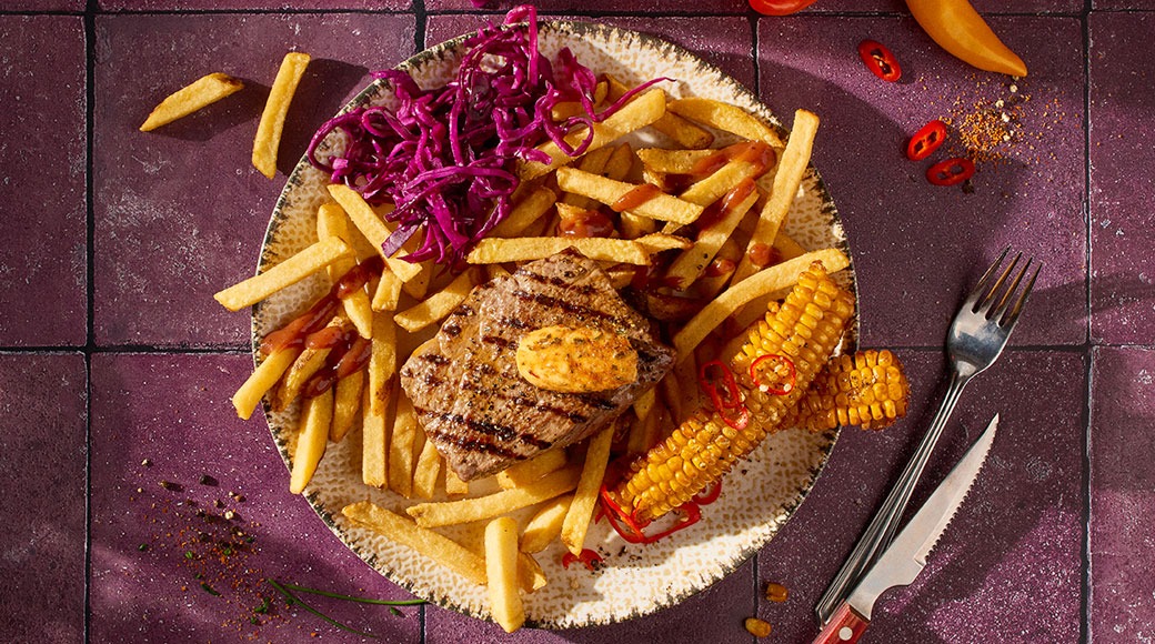 Grillattu pihvi voinokareella, ranskalaisia perunoita, punakaalisalaattia ja grillattua maissia lautasella. Vieressä haarukka ja veitsi violetilla laatoitetulla pöydällä.
