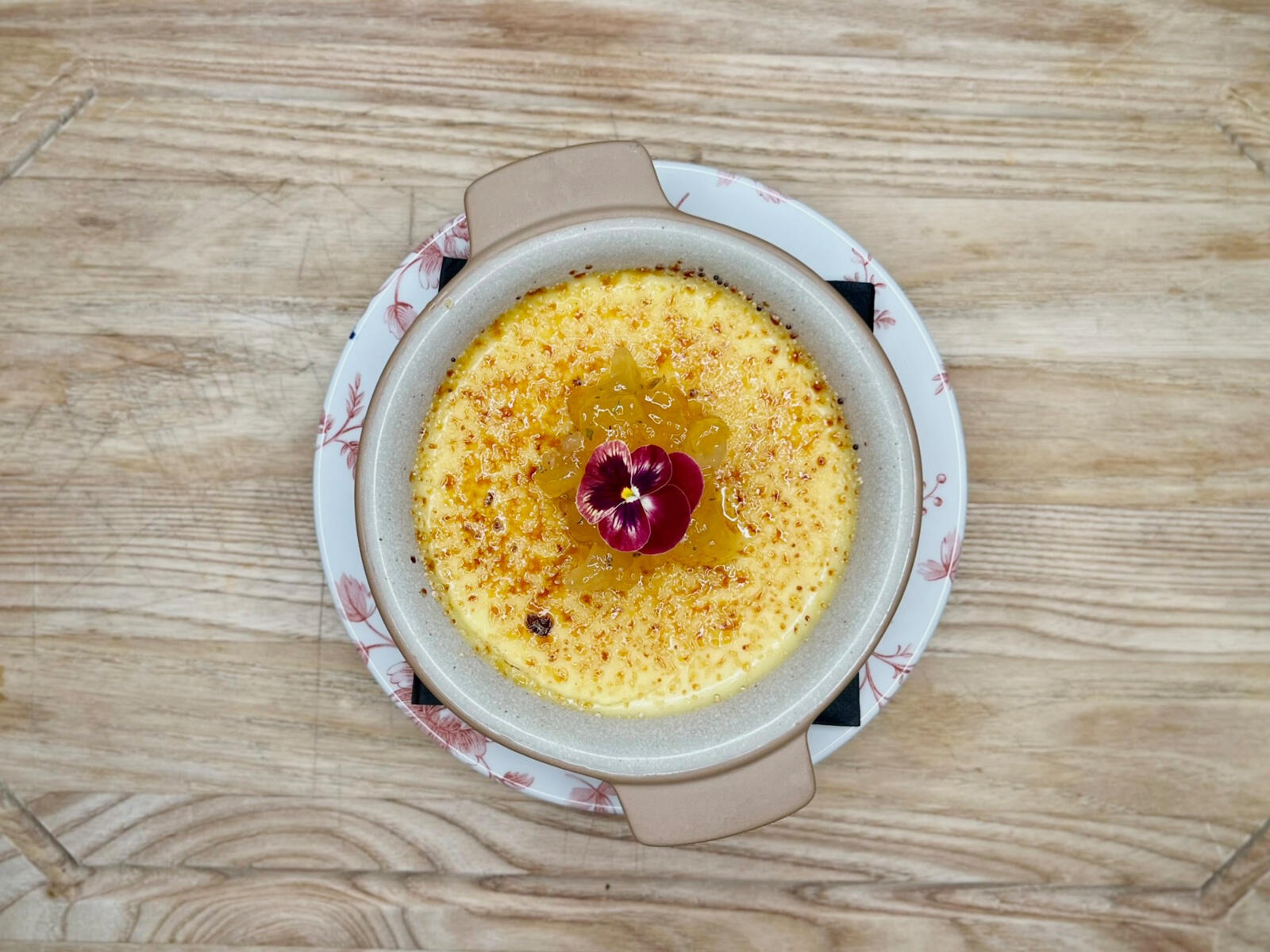 Crème brûlée in a ceramic dish topped with jam-like sauce and a purple edible flower, on a wooden table.