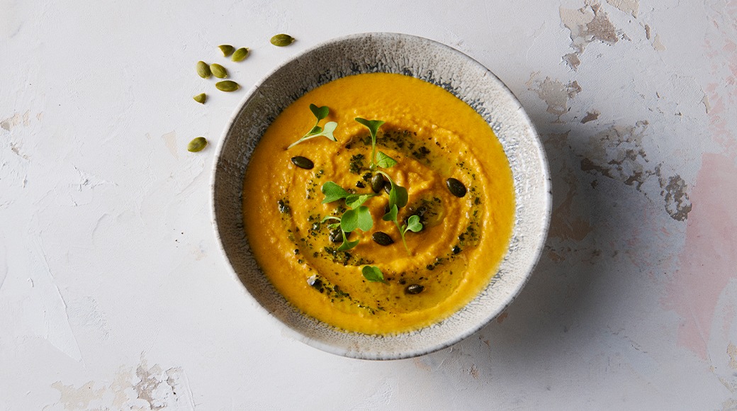 Orange pumpkin soup in a ceramic bowl, topped with herb oil, pumpkin seeds and sprouts. A few seeds lie on the table.