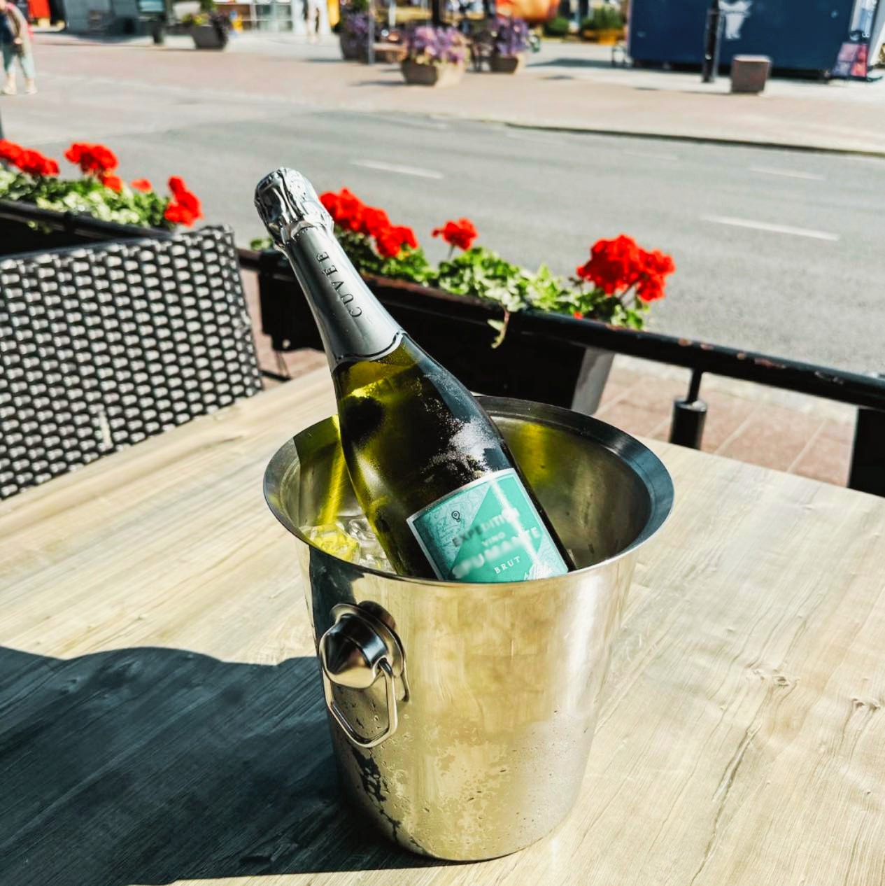 Kuohuviinipullo jäähdytysastiassa ravintolan terassilla, taustalla katu ja punaisia kukkia.