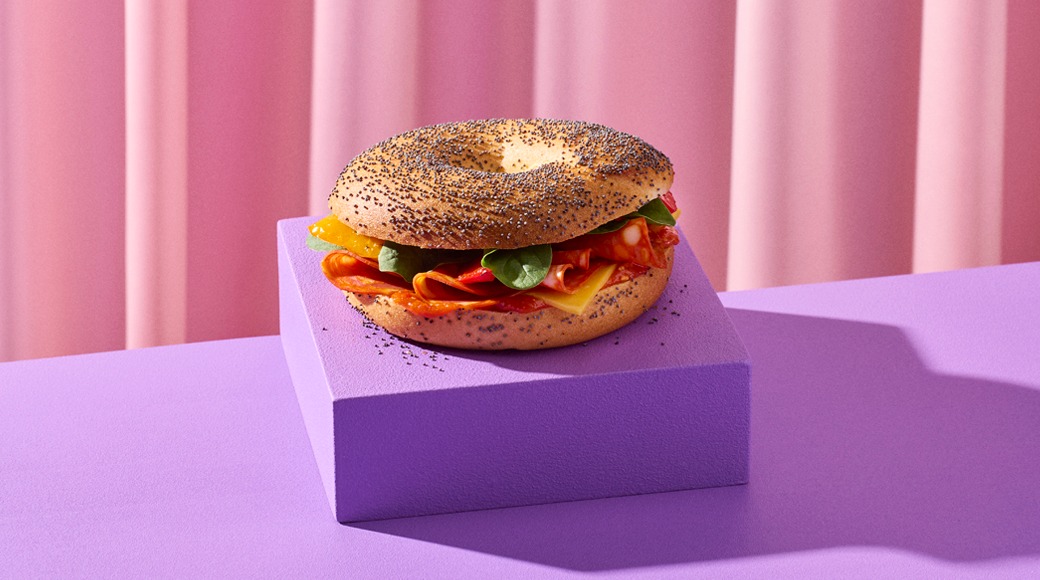 Poppy seed bagel filled with salami, cheese and spinach leaves. It sits on a purple block in front of a pink draped backdrop.