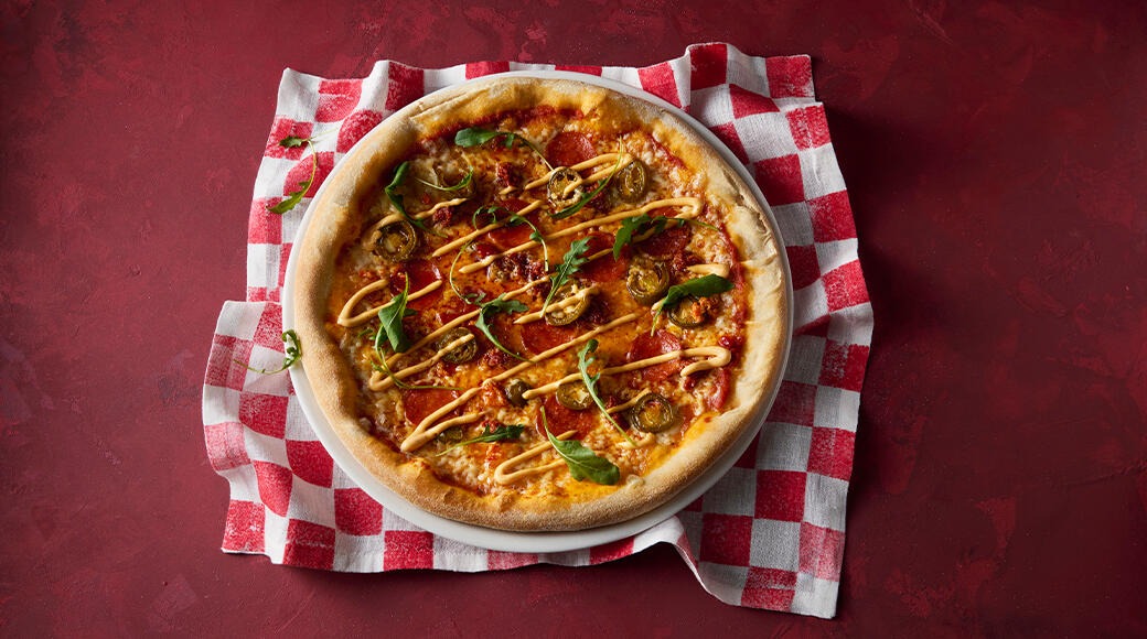 Pizza topped with pepperoni, jalapeños, rocket and a drizzle of creamy sauce. It sits on a white plate on a red and white checkered cloth.