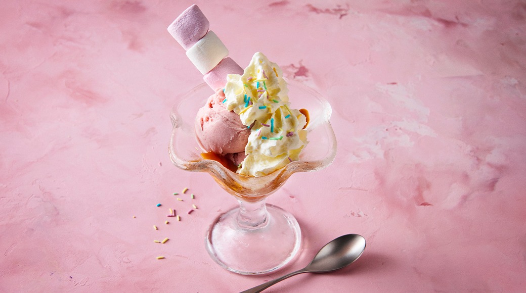 Ice cream sundae in a glass bowl with pink ice cream, whipped cream, colourful sprinkles and marshmallows. A spoon lies beside it on a pink background.