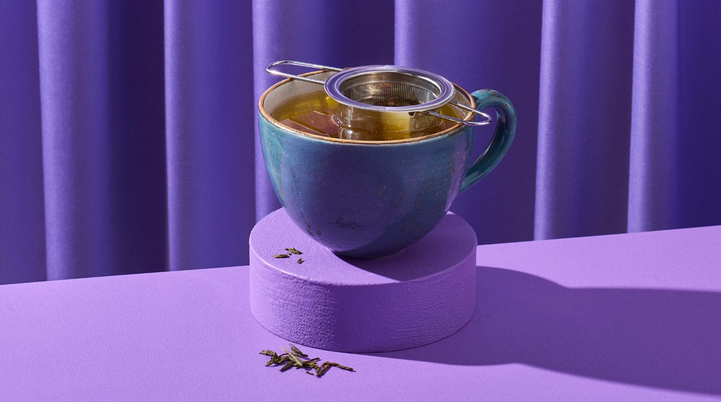 Sinertävä keraaminen teekuppi violetilla korokkeella. Kupin päällä on metallinen teesiivilä, ja ympärillä on teelehtiä. Taustalla on violetti verho.