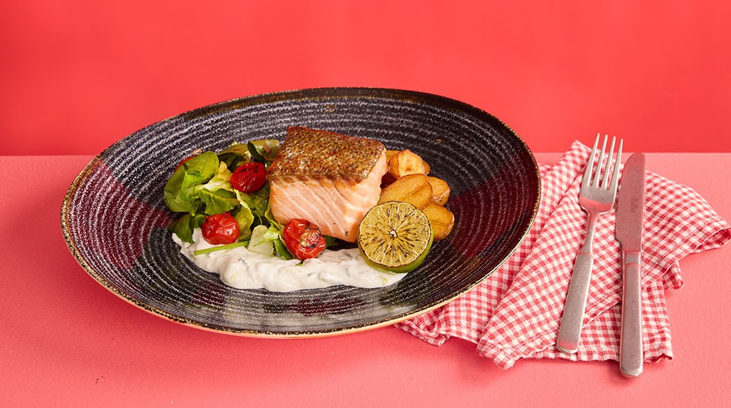 Lõhefileetükk tumedal taldrikul rohelise salatiga, tomatite, kartuliviilud ja laimimuna roosal laual.