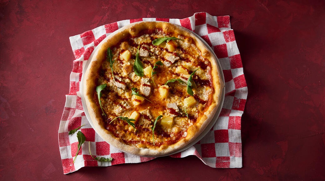 Pizza topped with chicken, pineapple, cheese and arugula. The pizza sits on a red and white checkered cloth on a red table.