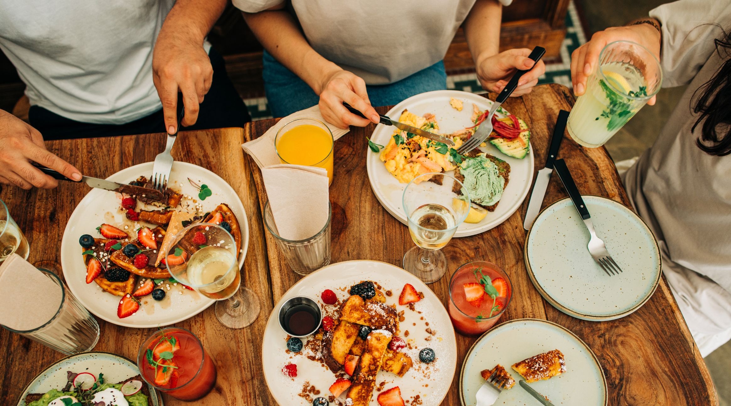 Useat henkilöt nauttivat aamiaisesta puisella pöydällä. Lautasilla on vohveleita, hedelmiä, munakokkelia ja avokadoleipää. Juomina mehua ja kahvia.