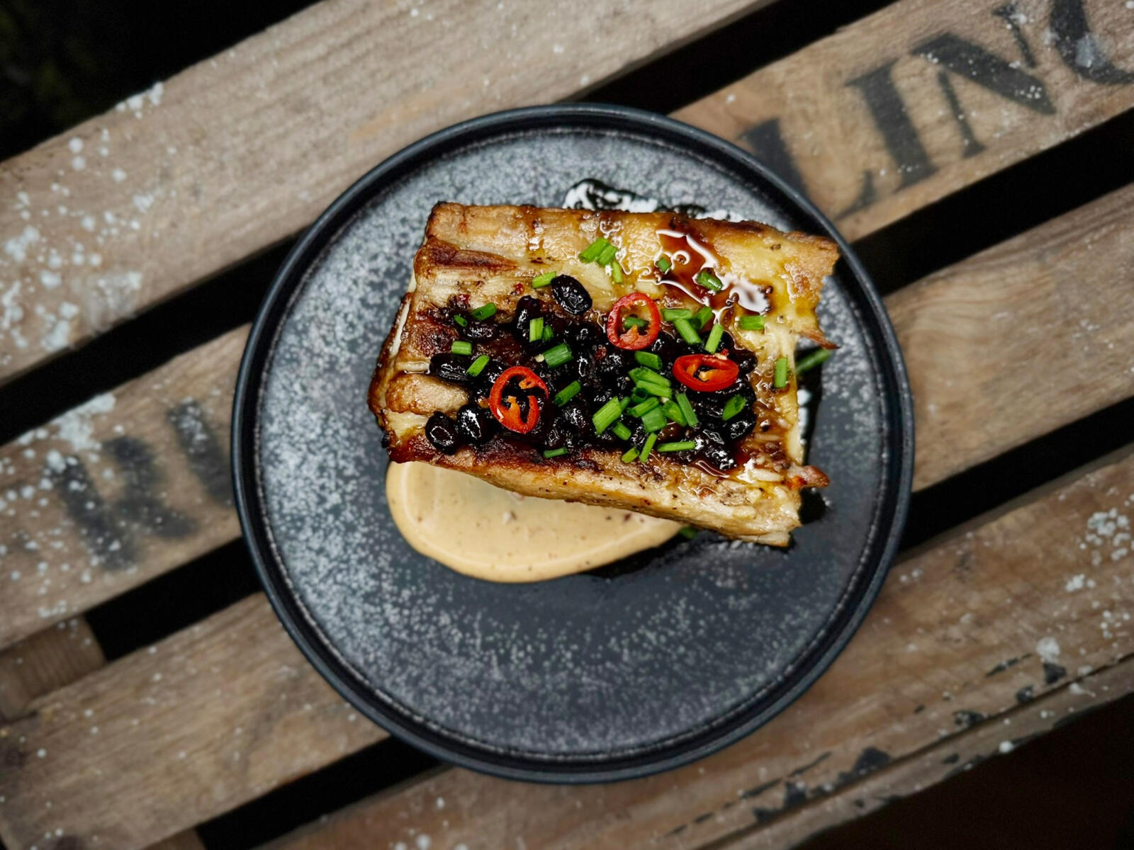 A piece of meat served on a black plate, garnished with black beans, green onions and fresh chili slices, with a creamy sauce underneath.