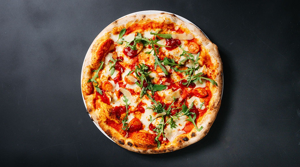 Pizza, jossa on tomaattikastiketta, juustoa, rucolasuikaleita ja kirsikkatomaatteja, tummalla alustalla.