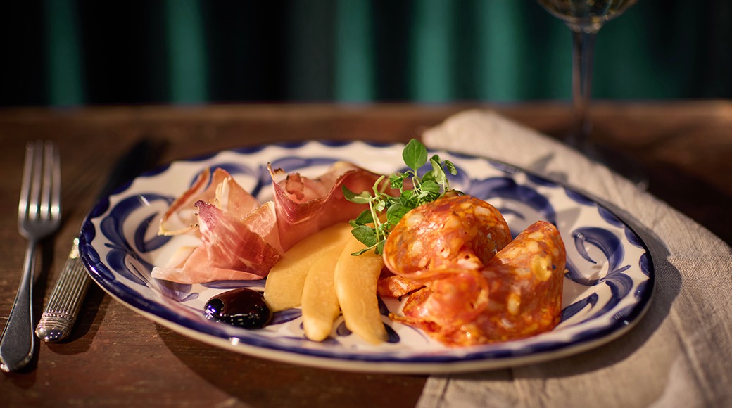 Spanska tapas på en tallrik: lufttorkad skinka, melonbitar och chorizokorv, garnerad med örter. Tallrik med blå-vit kant på en bordsduk.