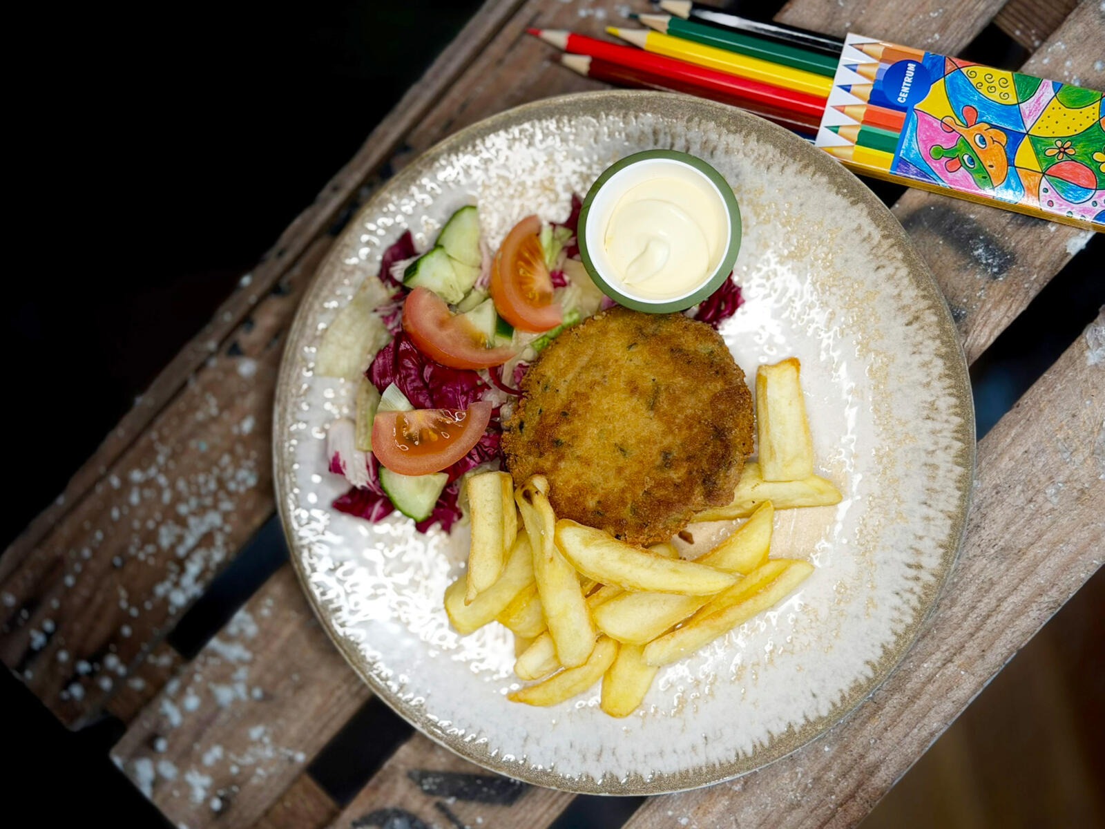 A plate with vegetable patty, french fries, salad and dip sauce. Colored pencils and a colorfully patterned box beside it.
