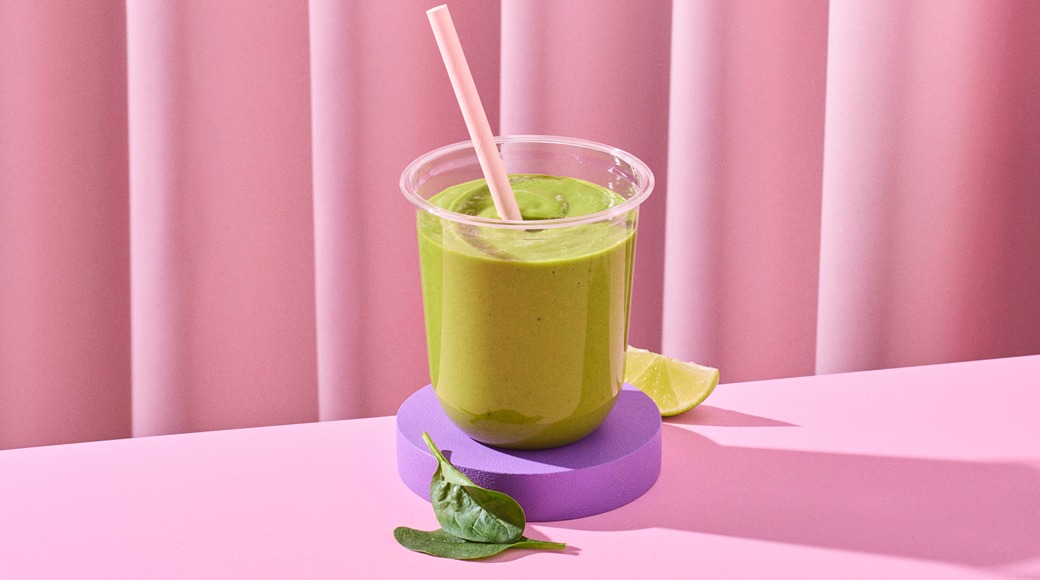 Vihreä smoothie kirkkaassa muovimukissa ja pinkissä pillissä violetin alustan päällä. Vieressä limelohko ja pinaatinlehtiä vaaleanpunaista taustaa vasten.