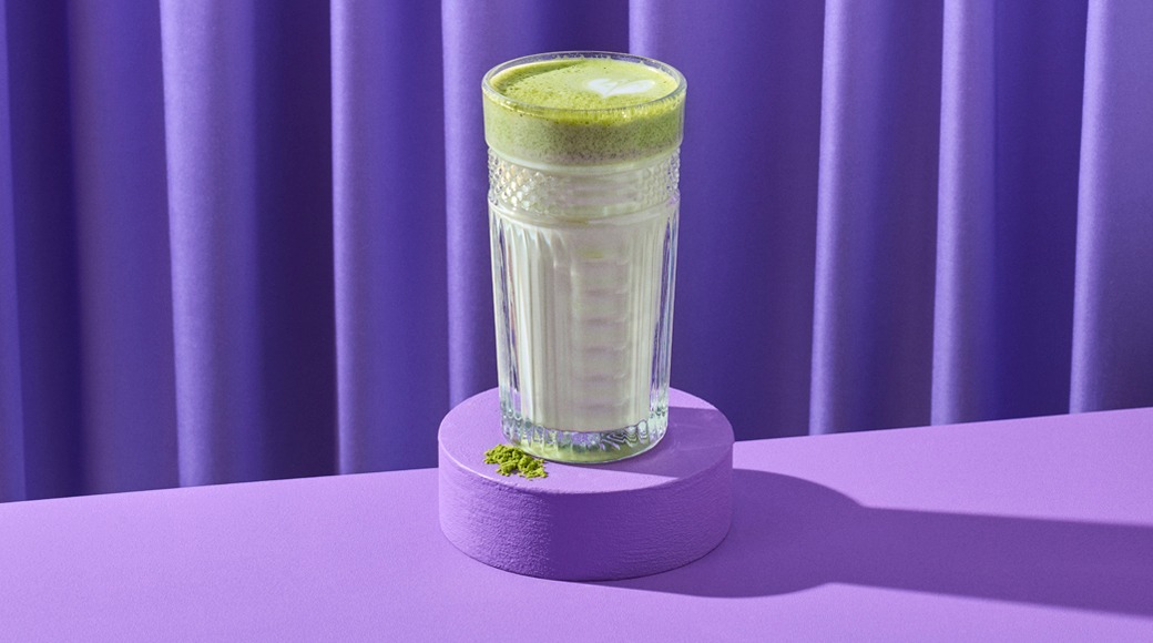 Matcha latte kirkkaassa kuvioidussa lasissa violetilla korokkeella. Lasin vieressä on vihreää matcha-jauhetta, taustalla violetti verho.