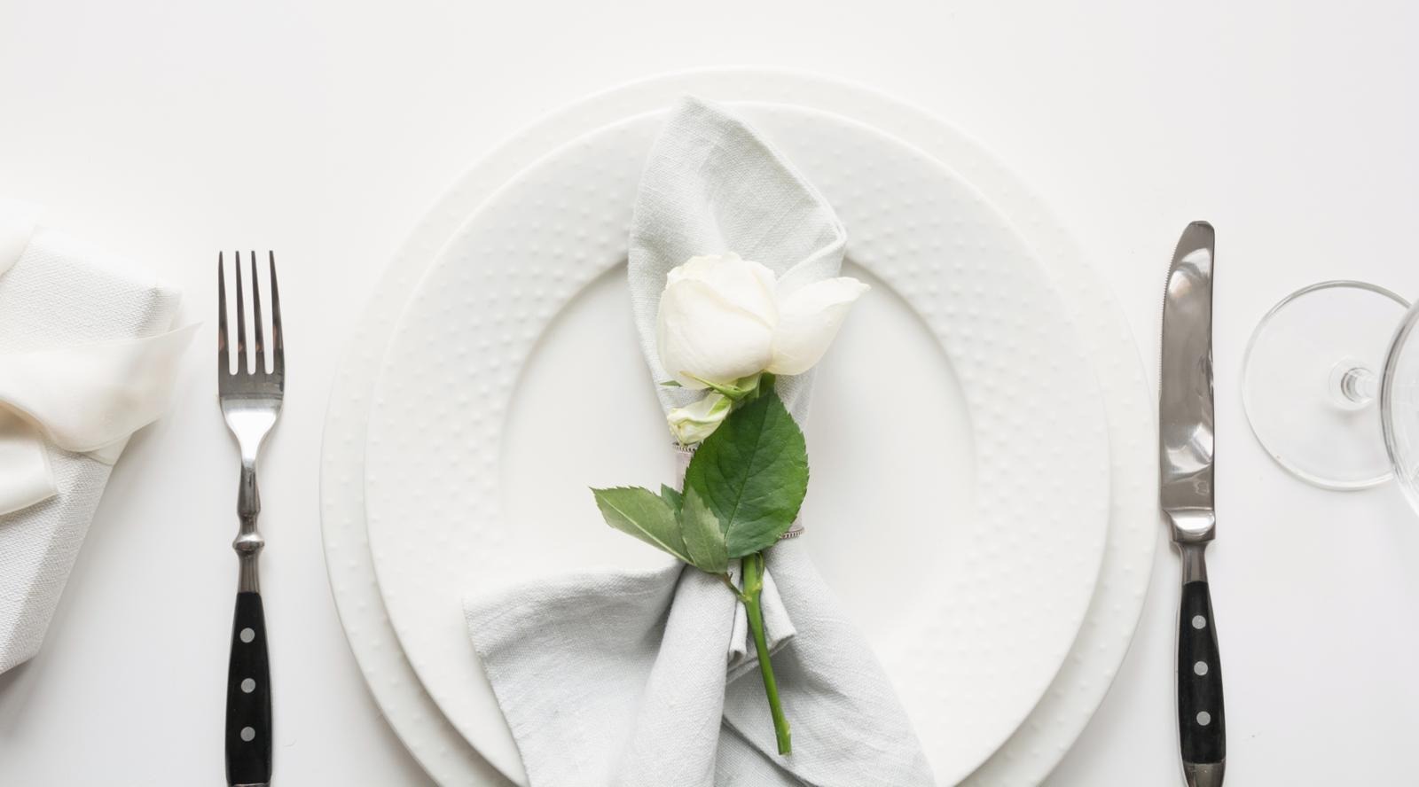 Valkoinen ruusu valkoisella lautasella valkoisella pöydällä. Vieressä aterimet.