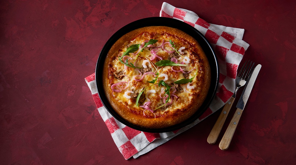 Cheesy pizza topped with red onion, rocket and shrimp on a black pan, resting on a red and white checked cloth. A fork and knife lie beside it.