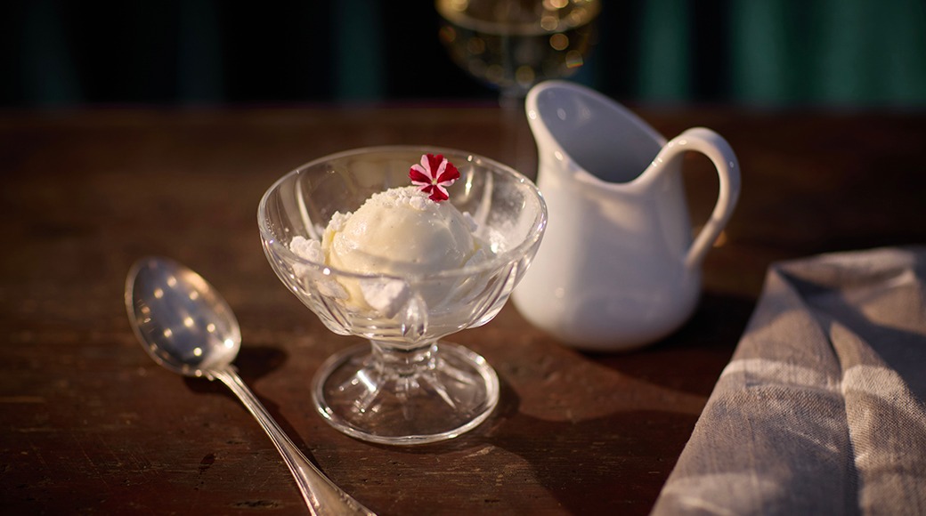 Klaasist magustoidukauss valge jäätisekuuliga, mida kaunistab väike punane-valge komm, kõrval hõbelusikas ja valge kann puidust laual.