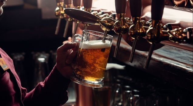 En hand fyller ett ölglas från en kran vid bardisken, med glänsande ölkranar i bakgrunden.