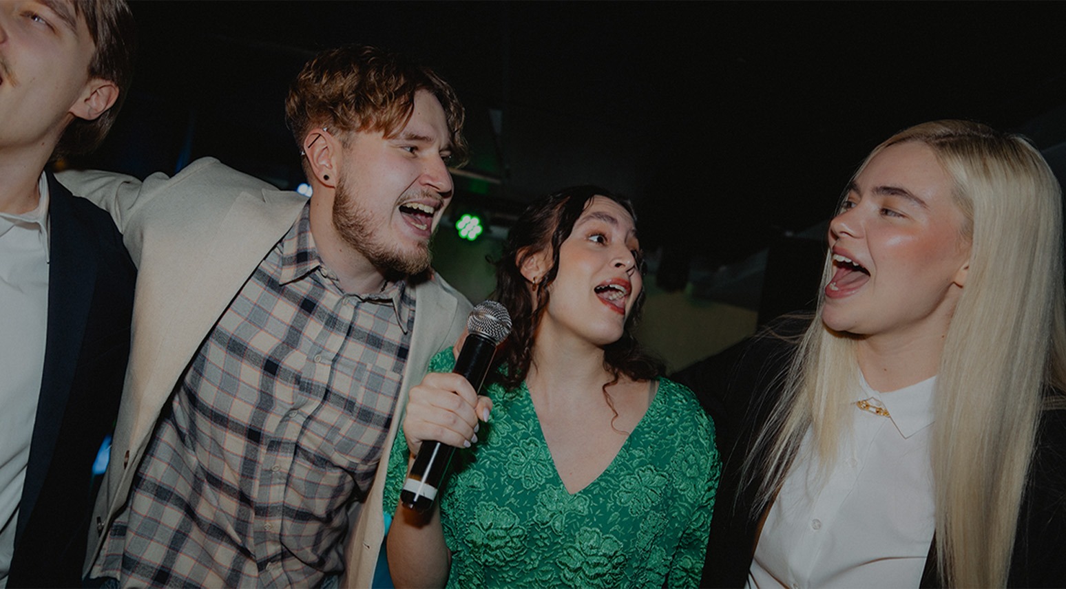Fyra personer sjunger tillsammans karaoke, en av dem håller en mikrofon. Belysning syns i bakgrunden.