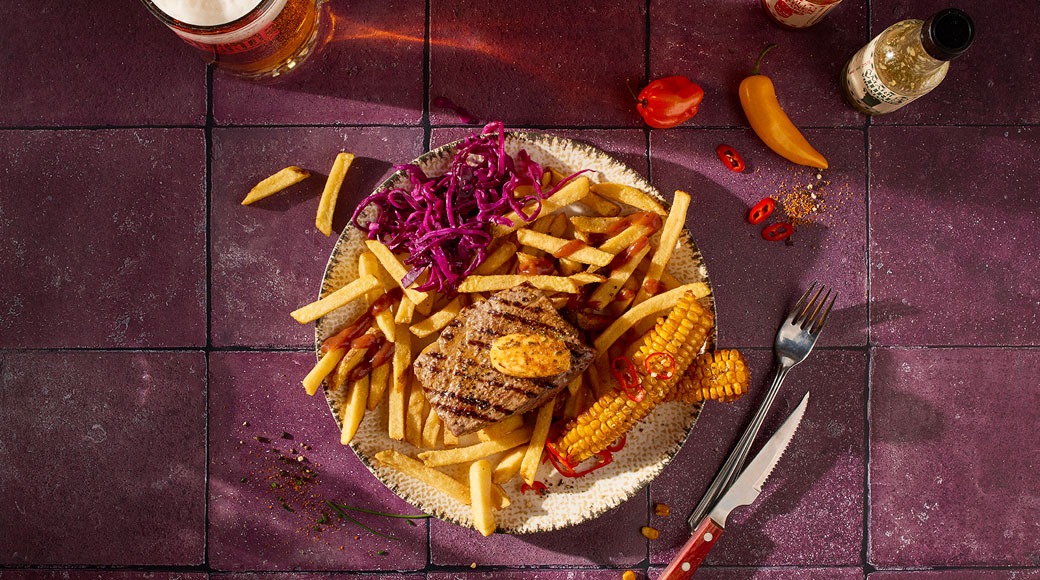 Pilt ülalt: grillitud praad võiga, friikartulid, punase kapsa salat ja mais taldrikul. Kõrval on kahvel, nuga, õlu ja tšillid lillal plaadil.
