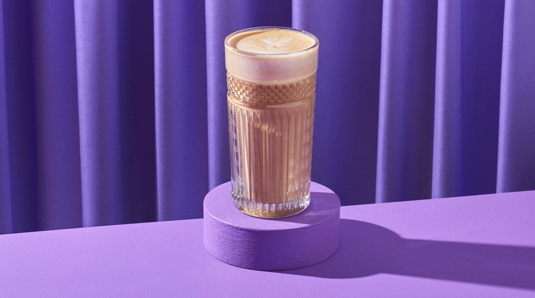 Kahvijuoma koristellussa lasissa violetilla korokkeella, taustalla violetti verho. Juoman päällä on latte-taidekuvio.