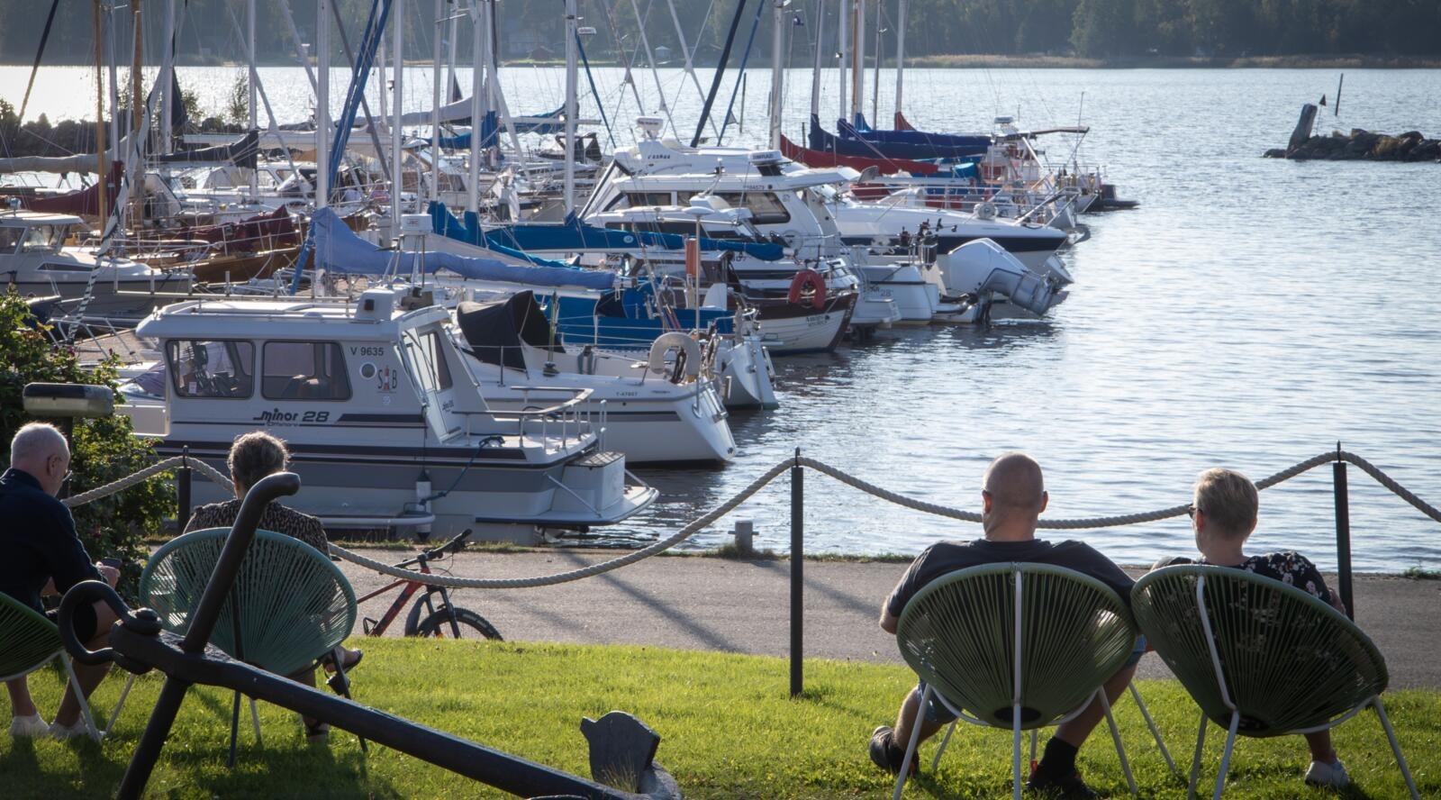 Personer sitter i stolar på gräsmattan och betraktar båtarna i hamnen, med en glittrande sjö i bakgrunden.