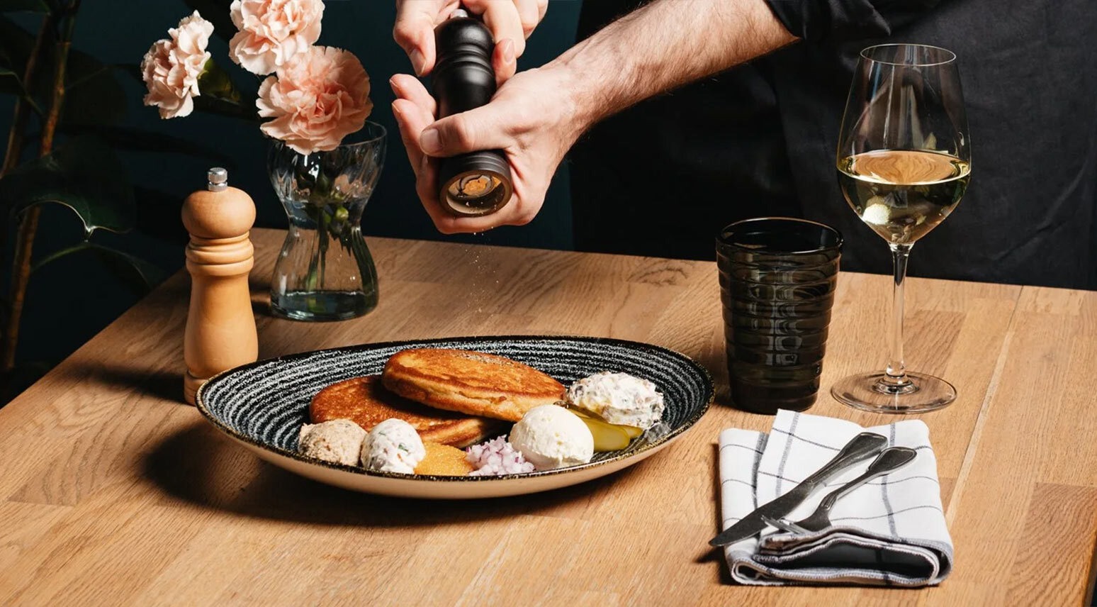 Henkilö jauhaa pippuria tyylikkäälle ruoka-annokselle puisella pöydällä. Pöydällä on valkoviinilasi, kukkia ja ruokailuvälineet.