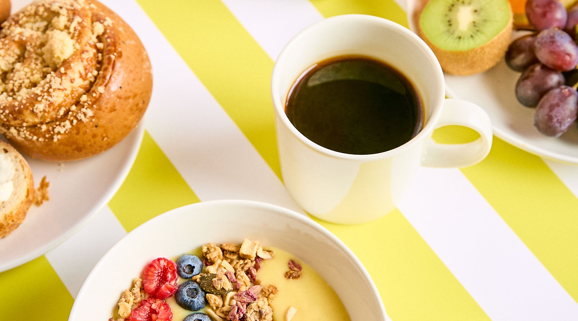 Breakfast table with yellow-striped tablecloth: cup of coffee, cinnamon roll, yogurt with berries and granola, and kiwi and grapes.