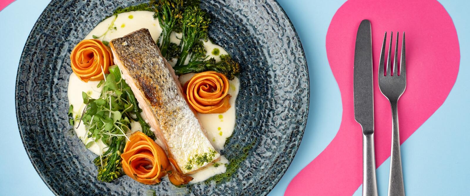 Stekt lax på en blå tallrik med krämig sås, dekorerad med morotsrosor och gröna örter.