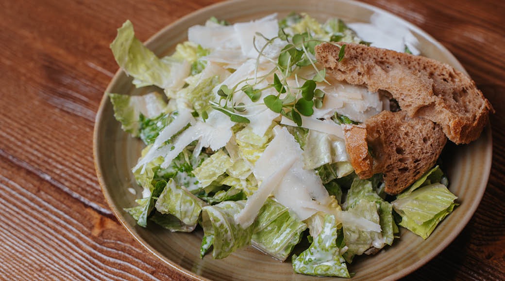 Caesar-salaatti keramiikka-astiassa, jossa parmesaanilastuja, salaattia, mikroyrttejä ja pala ruskeaa leipää puupöydällä.