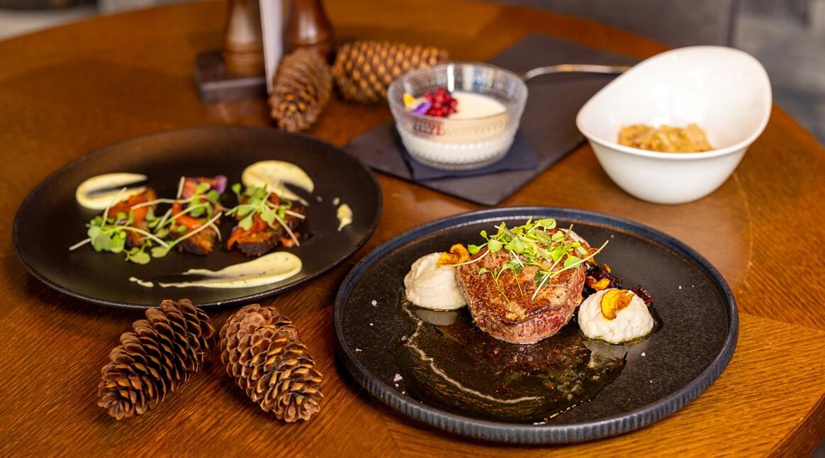 Round wooden table with two black plates of main courses, two bowls of desserts, and decorative pine cones.