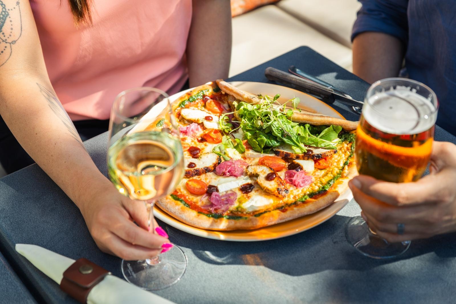 En pizza på tallrik, en person håller ett vinglas och en annan håller öl. Pizzan är toppad med grönsaker och kött.