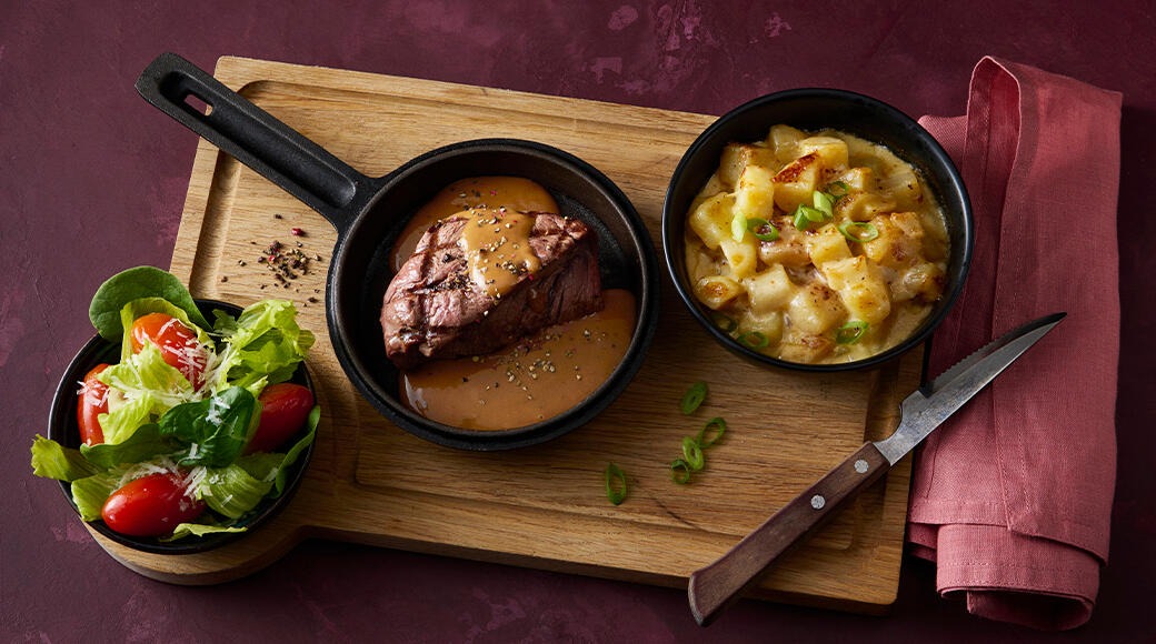 Grilled steak with sauce in a small cast-iron pan, beside creamy potatoes and a salad on a wooden board. A knife and a red napkin lie on the table.