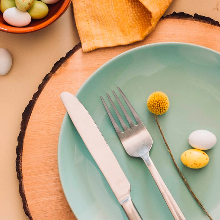 Turkoosi lautanen puisella alustalla, jossa on haarukka, veitsi, keltainen kukka ja pieniä pääsiäismunia.