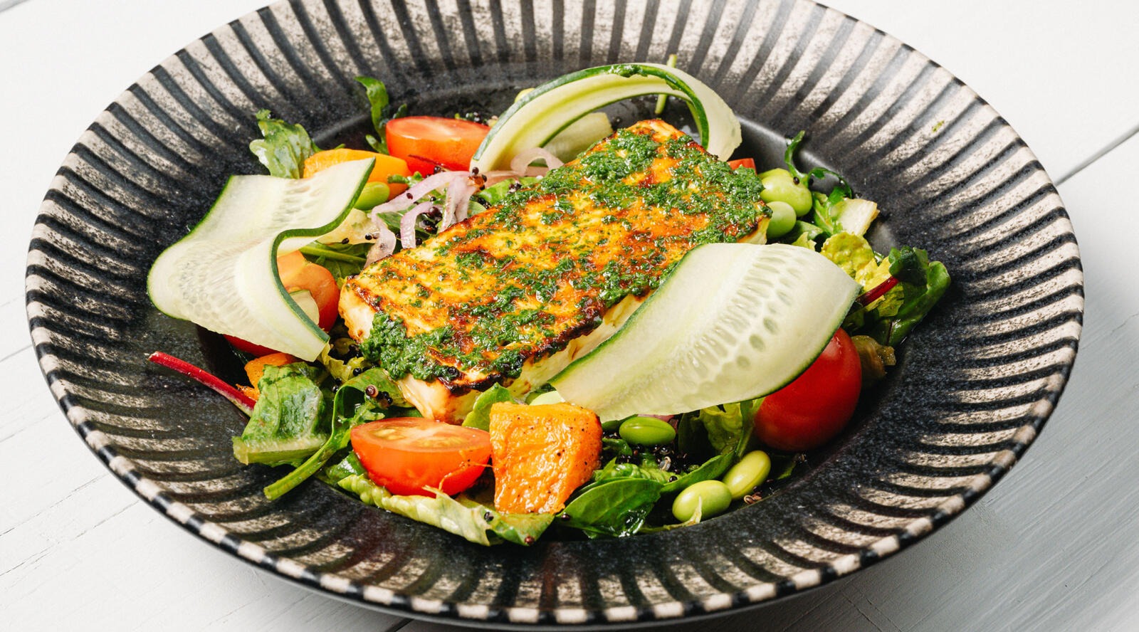 Salad on a plate with grilled fish fillet topped with green sauce, tomatoes, cucumber and beans.