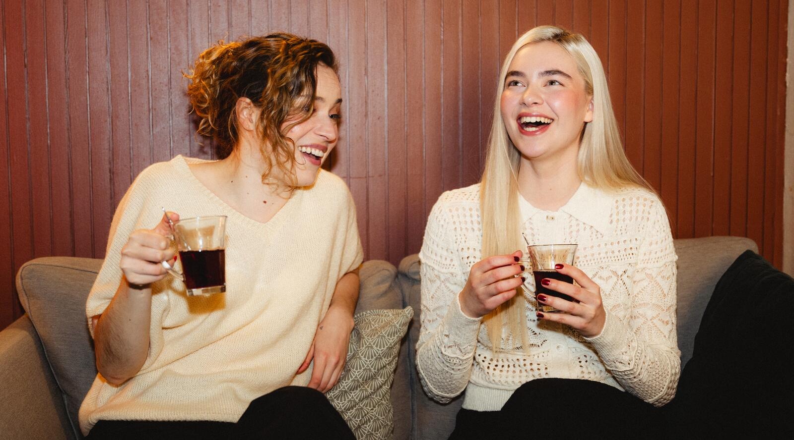 Två leende kvinnor sitter sida vid sida på en soffa och håller dryckesglas i sina händer.