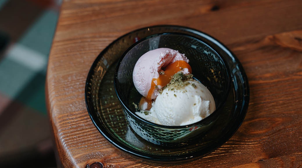 Jäätelöannos tummassa lasikulhossa puisella pöydällä. Valkoista ja vaaleanpunaista jäätelöä karamellisiirapilla ja vihreällä ripotteella.