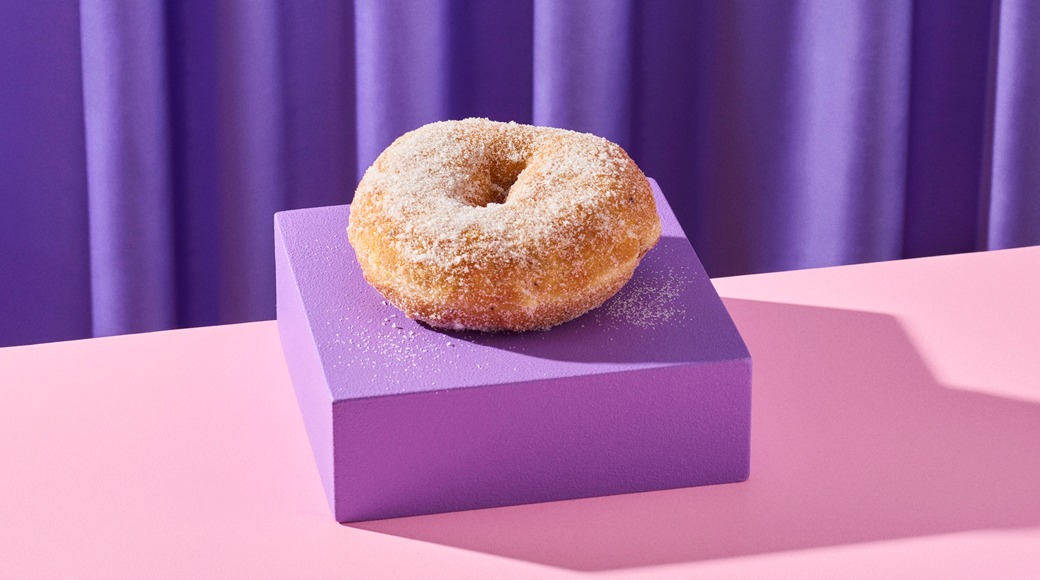 Sokerilla kuorrutettu donitsi violetilla korokkeella vaaleanpunaisella pinnalla, taustalla violetti verho.