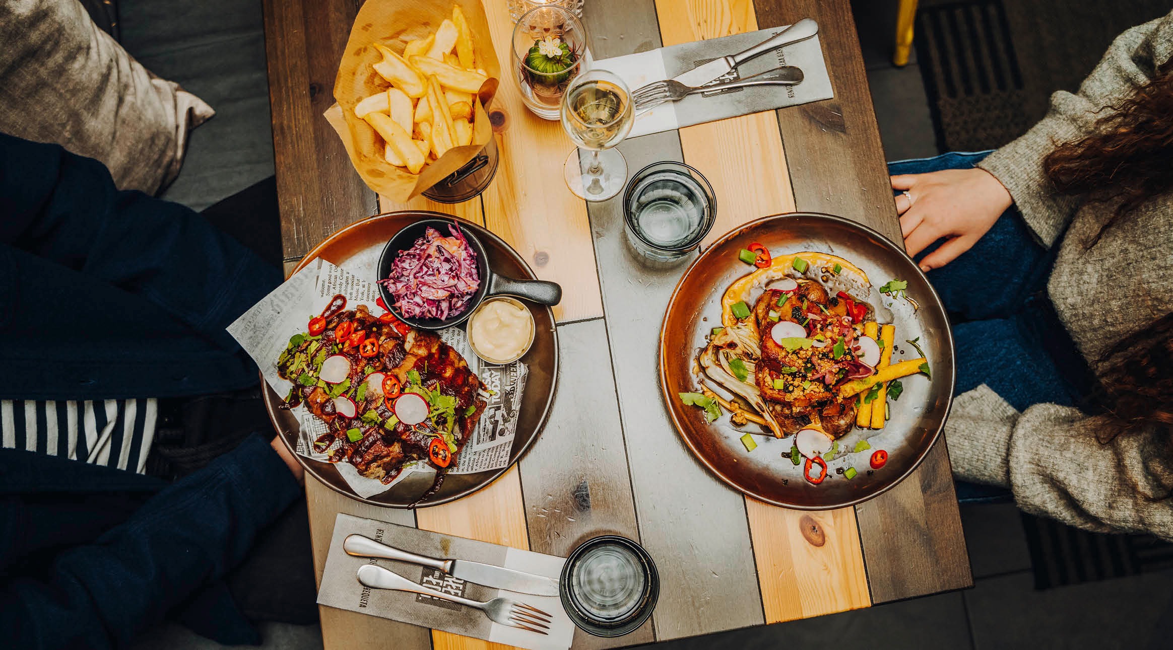 Näköala puupöydälle, jossa tarjoillaan kaksi värikkäästi koristeltua ruoka-annosta, ranskalaisia perunoita, punakaali-lisuketta, viinilasi ja vettä.