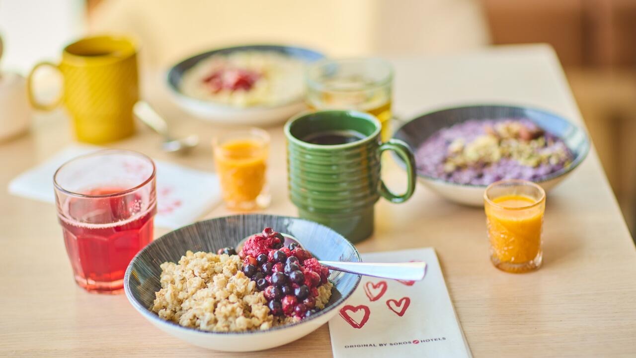 Aamiaispöytä, jossa on puuroa marjoilla, smoothiekulhoja, mehua, kahvia keltaisessa ja vihreässä mukissa sekä appelsiinimehua pienissä laseissa.