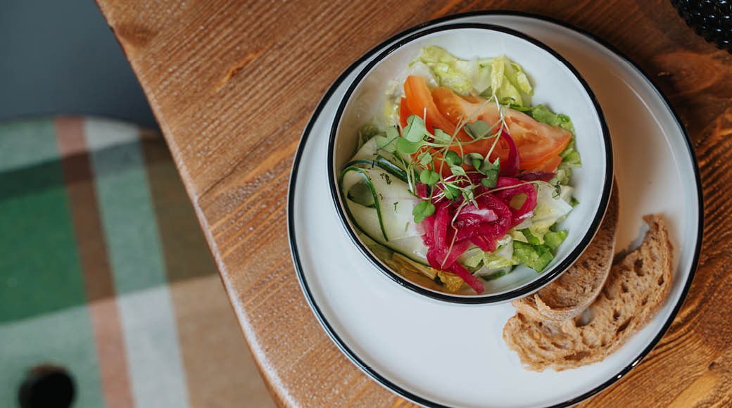 Värikkään salaatin annos valkoisessa kulhossa, tarjoiltuna leivän siivun kanssa puisella pöydällä.
