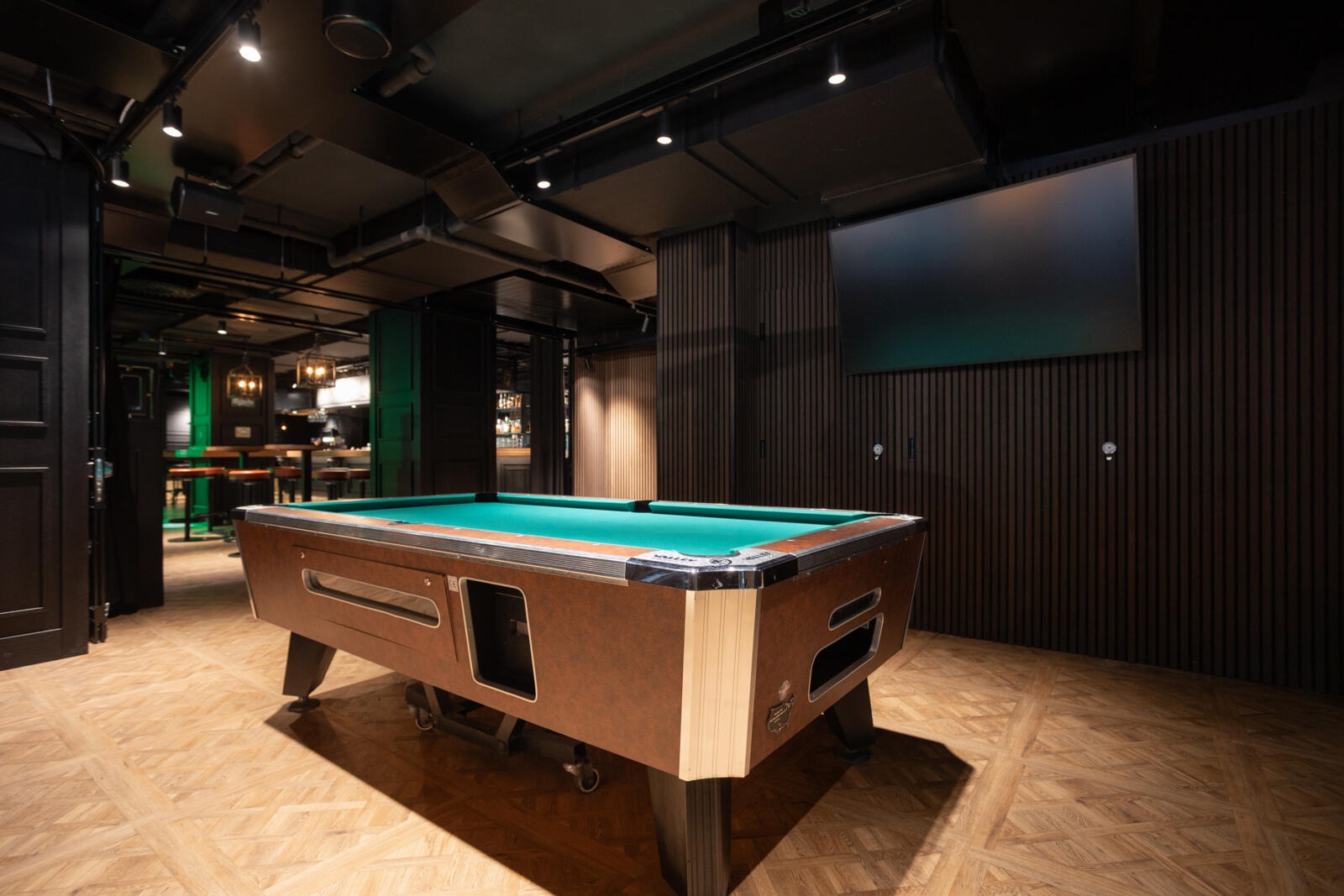 Biljardipöytä turkoosinvärisellä kankaalla tummassa baari- tai pelitilassa. Tilassa on puinen parkettilattia ja seinäpaneelit.